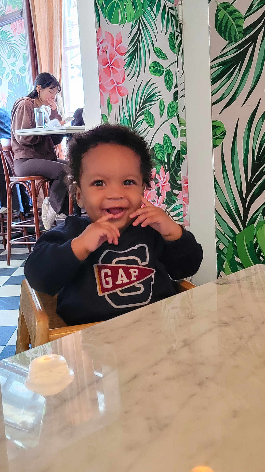 Lucas joined the competition — help win amazing prizes! baby, toddler, child, high_chair, smiling, person, indoor, table, marble_table, wallpaper, floral_pattern, plant_motif, cafe, woman, phone, drink, wooden_chair, curtain, checkered_floor, casual_clothing