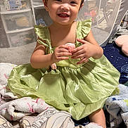 Kelly is registered to the contest to win money with this photo: toddler, child, smiling, green_dress, blanket, mesh_gate, indoor, happy, sitting, cute, baby, clothing, person, soft_toys, home, portrait, fabric, furniture, cozy, cute_face