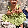 toddler, child, smiling, green_dress, blanket, mesh_gate, indoor, happy, sitting, cute, baby, clothing, person, soft_toys, home, portrait, fabric, furniture, cozy, cute_face