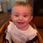 Eadie Rose joined the competition — help win amazing prizes! baby, child, smile, high_chair, indoors, clothing, person, happy, face, wooden_furniture, cute, young_child, portrait, seated, white_shirt, embroidery, straps, infant, blurred_background, eyes