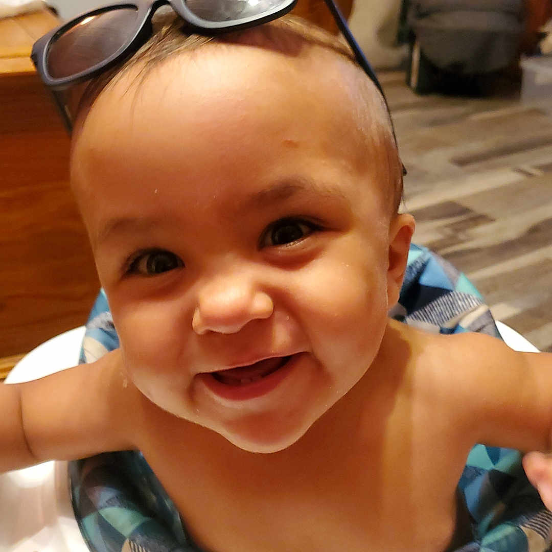 Grayson is registered to the contest to win money with this photo: baby, child, smiling, high_chair, sunglasses, indoor, face, happy, cute, infant, person, cheerful, headwear, young_child, wooden_floor, table, blue, skin, eyes, mouth