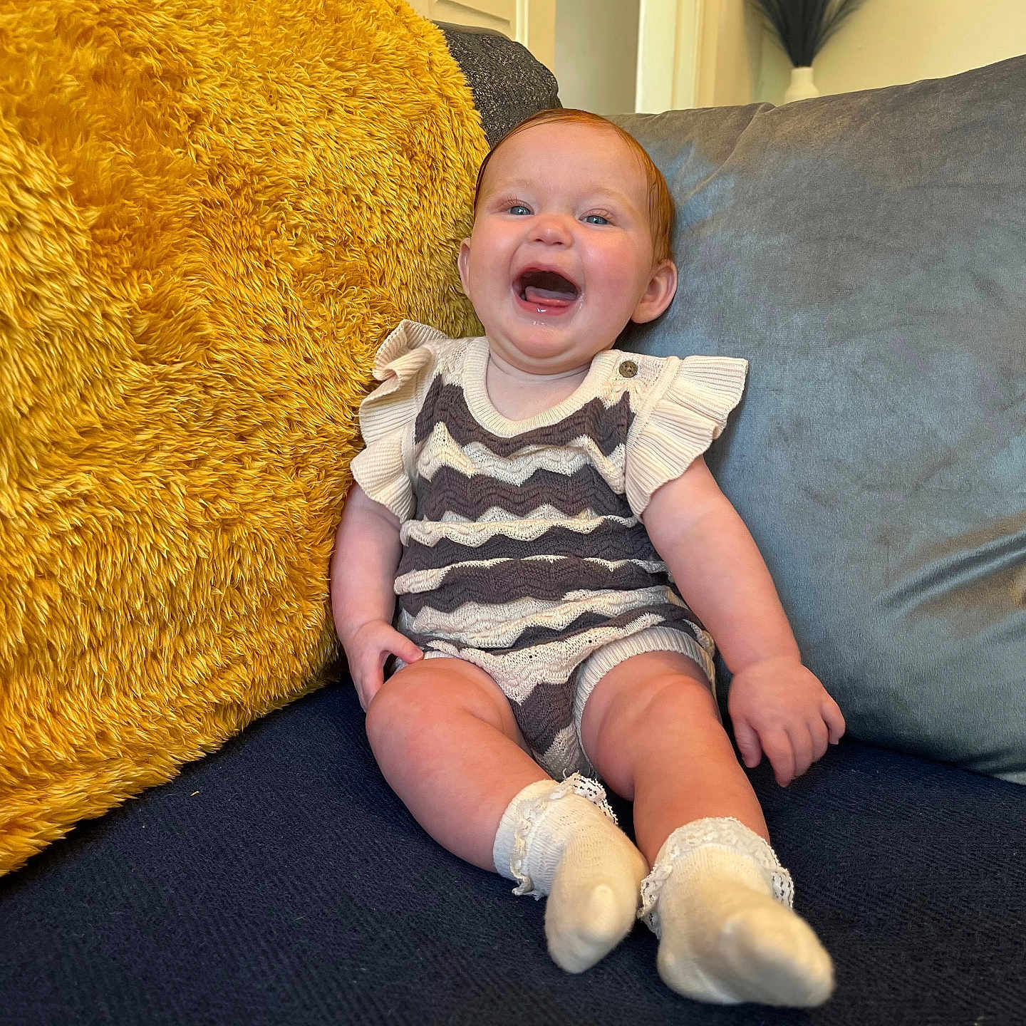 Esmae joined the competition — help win amazing prizes! baby, blue_couch, child, couch, cozy, cushion, cute, face, furniture, happy, head, home, indoor, infant, knitwear, person, sitting, smiling, sock, yellow_cushion