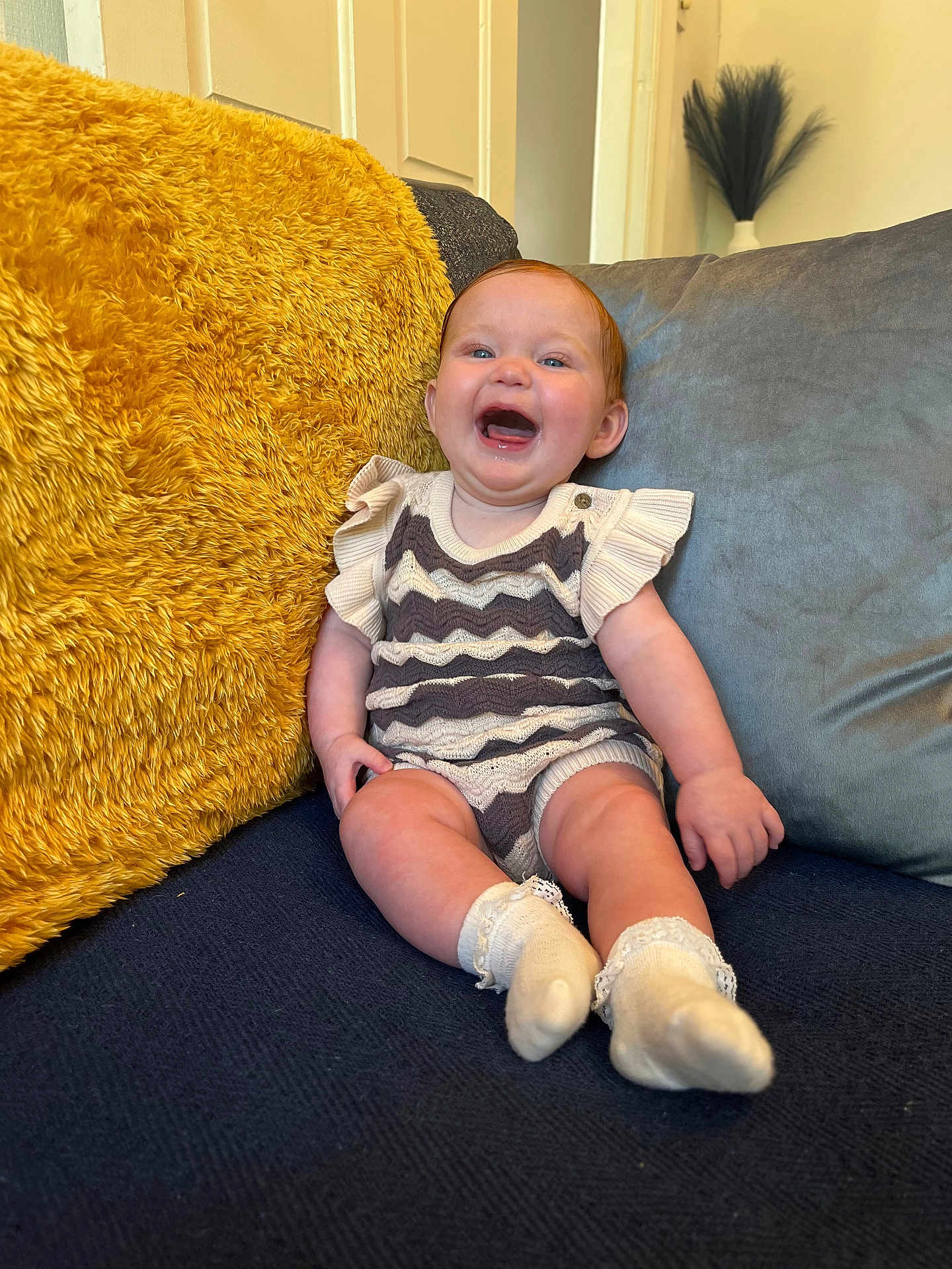 Esmae joined the competition — help win amazing prizes! baby, child, smiling, sitting, couch, cushion, yellow_cushion, blue_couch, happy, indoor, sock, knitwear, person, face, head, furniture, cozy, home, cute, infant