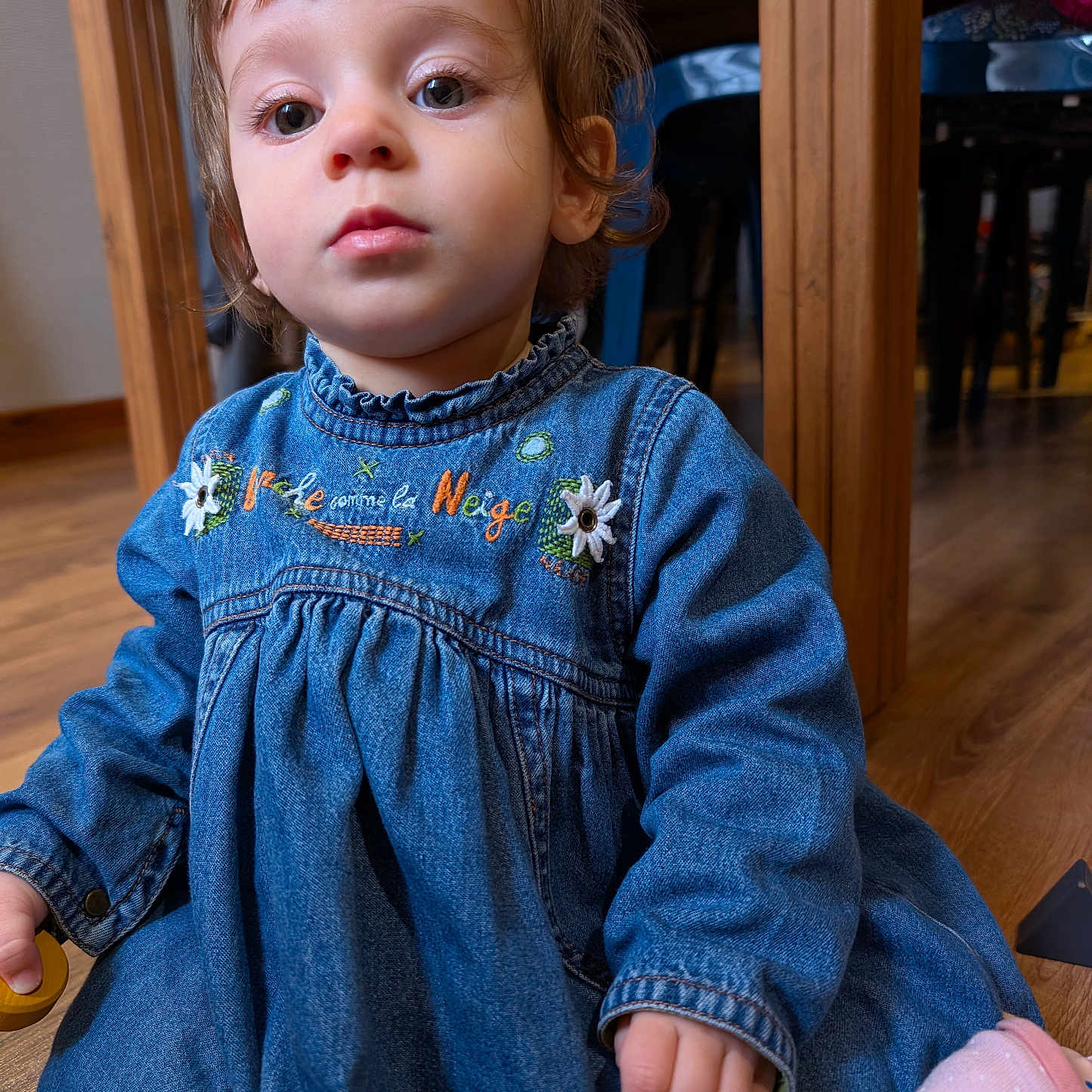 Alma participe au concours pour gagner de l'argent avec cette photo : big_eyes, blue_clothing, casual, chair, child, closeup, curly_hair, cute, denim_dress, face, floor, furniture, indoor, person, pink_toy, sitting, table, toddler, toy, wooden_floor