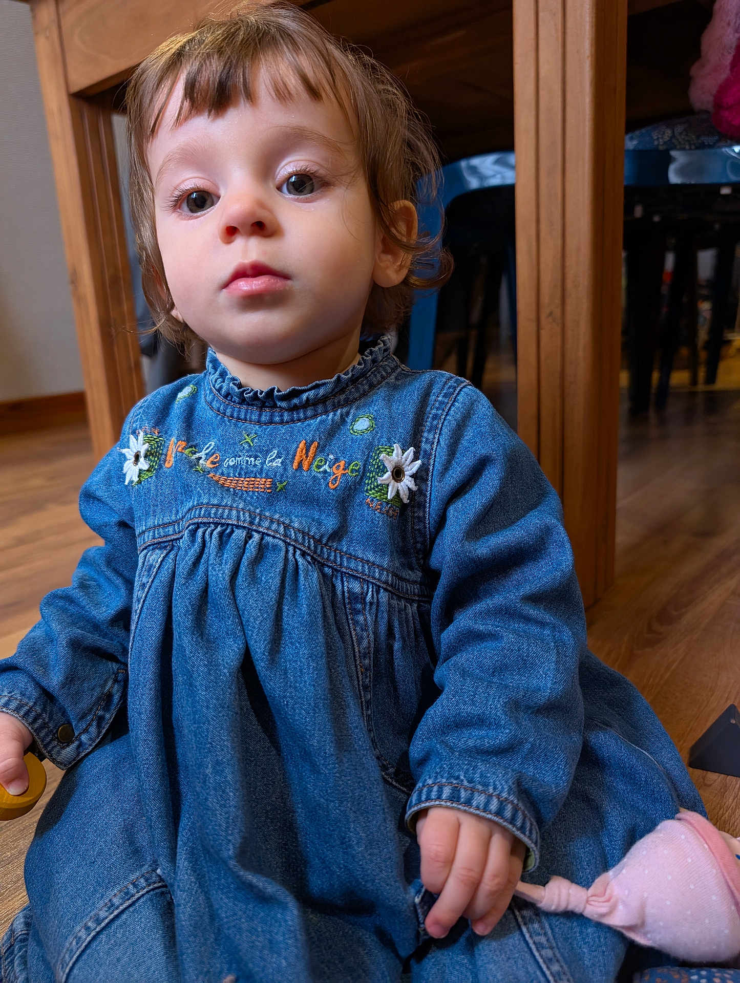 Alma participe au concours pour gagner de l'argent avec cette photo : toddler, child, denim_dress, indoor, wooden_floor, table, chair, toy, pink_toy, curly_hair, face, big_eyes, sitting, person, blue_clothing, floor, furniture, closeup, casual, cute