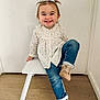 child, toddler, smiling, sitting, white_chair, blue_jeans, beige_shoes, pigtails, long_sleeve_shirt, wooden_floor, indoor, white_wall, cute, happy, casual_clothing, playful, young_child, portrait, small_chair, buttoned_shirt