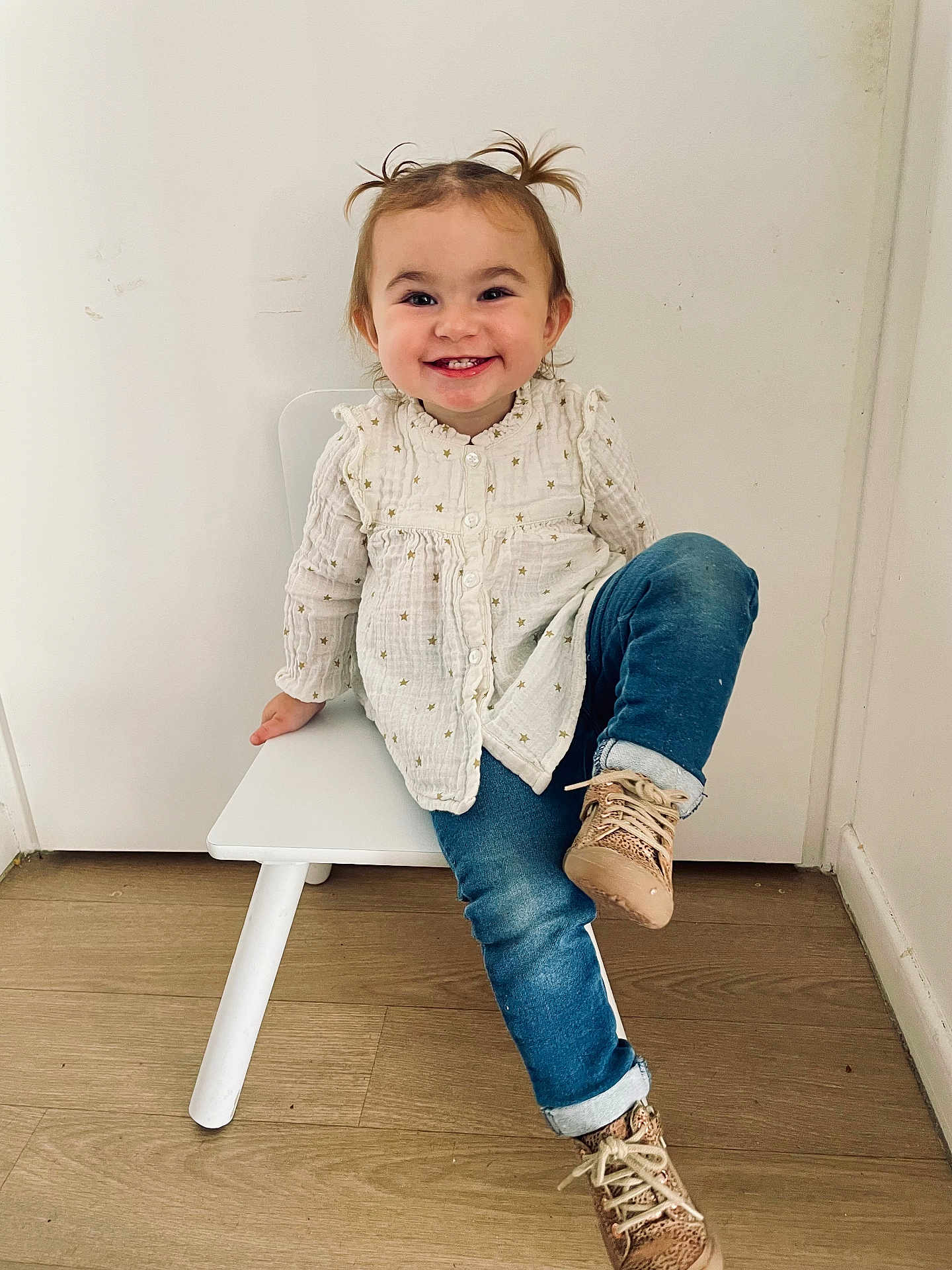 Elya a rejoint le concours — aidez-le/la à gagner de superbes lots ! child, toddler, smiling, sitting, white_chair, blue_jeans, beige_shoes, pigtails, long_sleeve_shirt, wooden_floor, indoor, white_wall, cute, happy, casual_clothing, playful, young_child, portrait, small_chair, buttoned_shirt