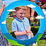 child, boy, smiling, blue_shirt, outdoor, sunny, grass, people, tattoos, hand_clasped, circular_frame, clouds, green_hills, casual_clothing, happy, portrait, daylight, playful_frame, event, park