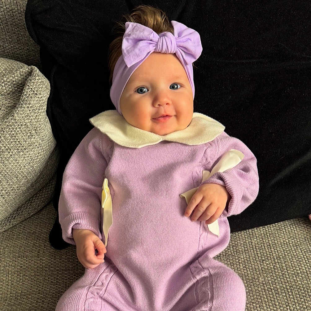 Monroe is registered to the contest to win money with this photo: baby, child, smiling, headband, bow, purple_outfit, sitting, couch, cushion, cozy, indoor, portrait, cute, happy, infant, clothing, hands, face, blue_eyes, soft_lighting