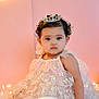 baby, child, tiara, dress, pink_background, balloon, flower_pattern, curly_hair, sitting, portrait, cute, indoors, soft_lighting, decorative_lights, serious_expression, young_child, celebration, birthday, adorable, fashion