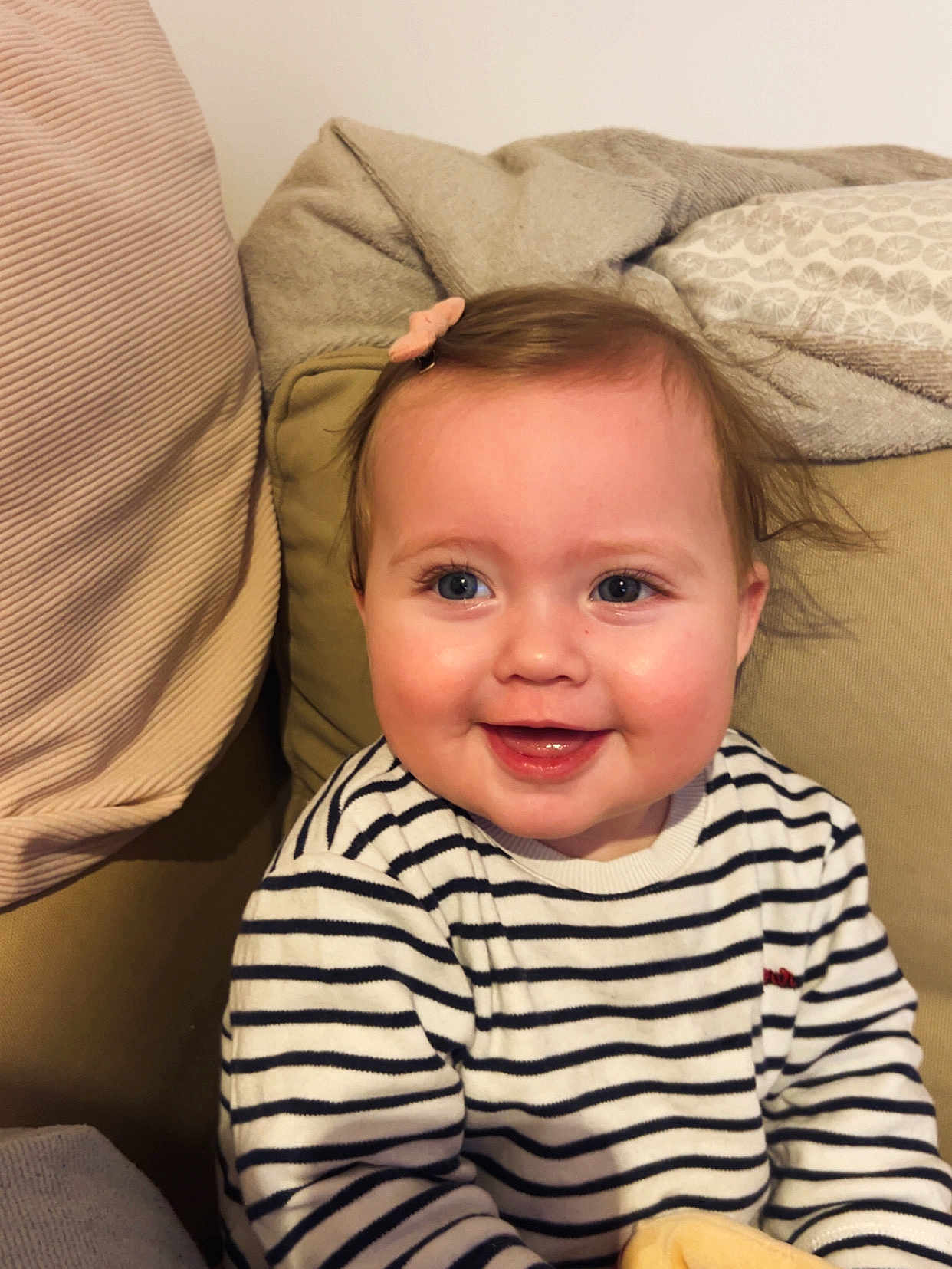 Giulia participe au concours pour gagner de l'argent avec cette photo : baby, child, smiling, striped_shirt, hair_clip, indoor, couch, blanket, happy, portrait, face, toddler, cozy, light_skin, blue_eyes, seated, clothing, cute, infant, home