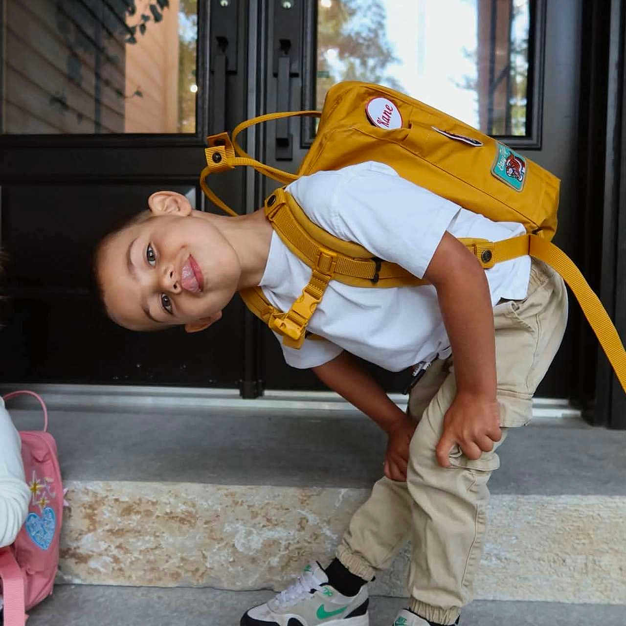 Kane joined the competition — help win amazing prizes! child, boy, backpack, yellow_backpack, outdoor, doorstep, portrait, standing, bending, playful, smiling, sneakers, white_shirt, beige_pants, school, travel, entrance, house, daylight, reflection