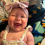 Averi is registered to the contest to win money with this photo: baby, blanket, bow, cheerful, child, closeup, clothing, cozy, cute, dress, face, happy, headband, indoors, infant, person, portrait, quilt, smiling, soft
