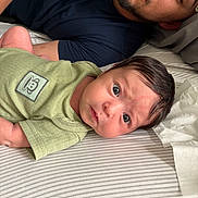 Brisa is registered to the contest to win money with this photo: baby, infant, man, beard, green_onesie, bed, striped_sheet, face, portrait, lying_down, indoor, person, curious, wide_eyes, head, expression, clothing, adult, skin, hair