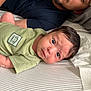 baby, infant, man, beard, green_onesie, bed, striped_sheet, face, portrait, lying_down, indoor, person, curious, wide_eyes, head, expression, clothing, adult, skin, hair