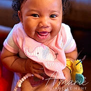 Amarie joined the competition — help win amazing prizes! baby, smiling, pink_bib, elephant_embroidery, toy, hand, happy, closeup, infant, cute, child, person, portrait, indoors, warm_lighting, cheerful, holding, clothing, cute_expression, adorable
