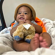 Eli is registered to the contest to win money with this photo: baby, bed, blanket, child, closeup, cozy, cute, feet, happy, hat, indoor, infant, knit_hat, orange_blanket, person, portrait, smiling, soft, teddy_bear, toy