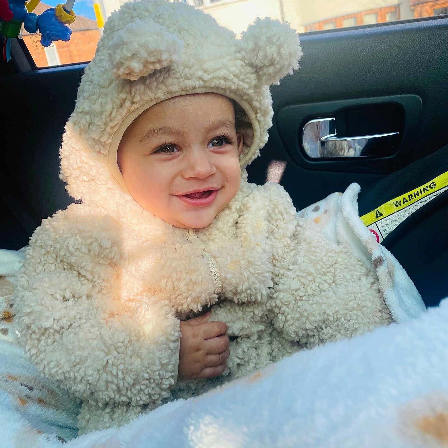 Ekam is registered to the contest to win money with this photo: baby, bear_costume, blanket, car_seat, child, cute, face, fluffy_clothing, hand, happy, indoor, infant, portrait, seatbelt, smiling, sunlight, toy, vehicle_interior, warm_clothing, window