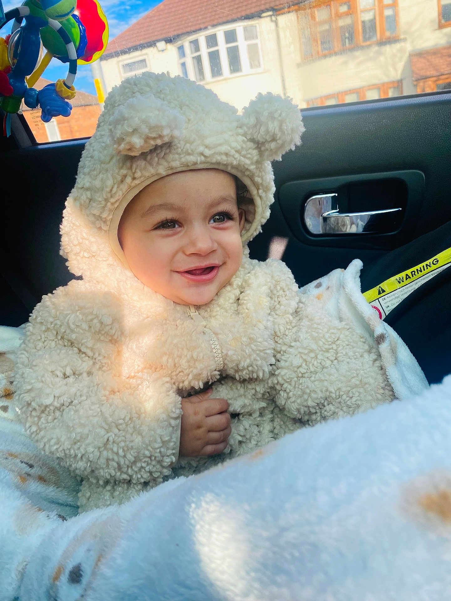 Ekam is registered to the contest to win money with this photo: baby, child, smiling, cute, fluffy_clothing, bear_costume, car_seat, blanket, toy, window, sunlight, face, hand, indoor, seatbelt, vehicle_interior, happy, infant, warm_clothing, portrait