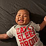 baby, smiling, lying_down, shirt, text, gray_clothing, black_background, happy, infant, cute, tongue_out, short_sleeves, child, person, skin, hair, indoors, toddler, playful, closeup