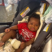 Timothy joined the competition — help win amazing prizes! baby, smiling, stroller, bottle, blanket, red_shirt, infant, indoor, happy, person, footwear, floor, clothing, curly_hair, baby_bottle, seat, child, cute, comfortable, relaxed