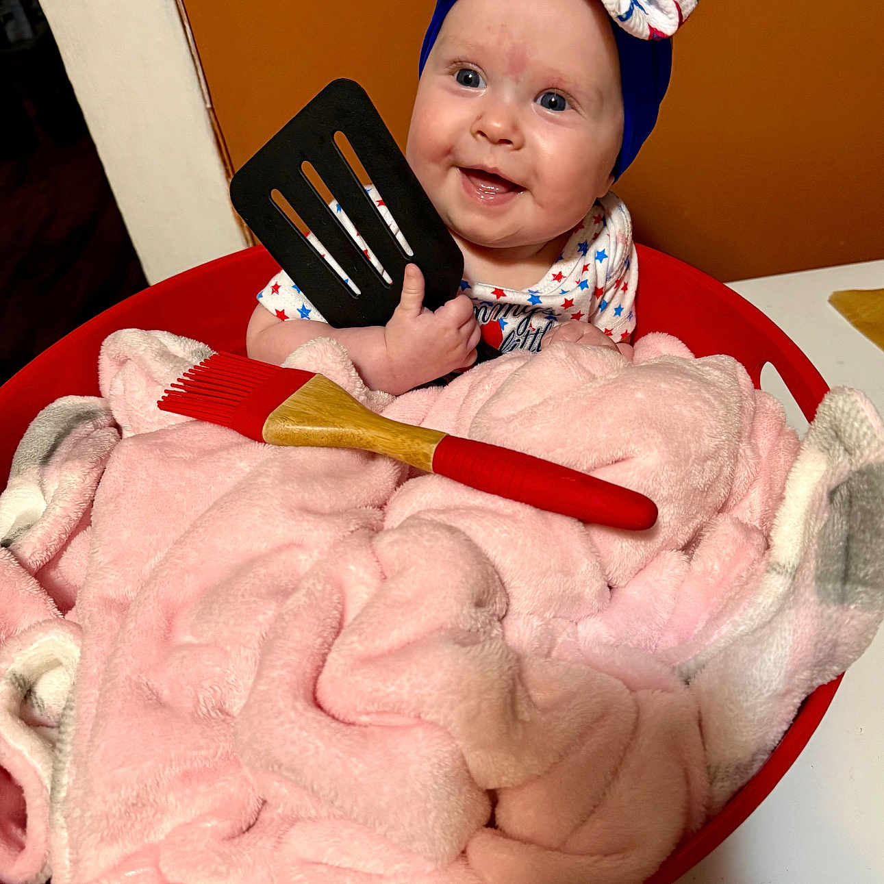 Brecklyn is registered to the contest to win money with this photo: baby, basin, blanket, bow, child, cozy, cute, happy, headwear, indoors, infant, kitchen_utensil, person, pink, playful, red, smiling, soft_texture, spatula, table