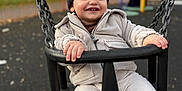 Myles is registered to the contest to win money with this photo: child, toddler, swing, playground, outdoor, hat, jacket, smiling, happy, chain, seat, autumn, trees, fence, play_equipment, person, park, daytime, clothing, fun