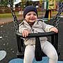 child, toddler, swing, playground, outdoor, hat, jacket, smiling, happy, chain, seat, autumn, trees, fence, play_equipment, person, park, daytime, clothing, fun