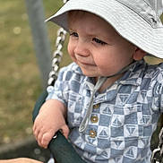 Myles is registered to the contest to win money with this photo: toddler, child, swing, hat, sunhat, shirt, patterned_clothing, hand, playground, outdoor, chains, focus, portrait, baby, expression, holding, support, daylight, grass, seat