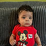 baby, child, mickey_mouse, red_shirt, curly_hair, couch, gray_couch, portrait, indoors, cute, infant, sitting, clothing, expression, hand, face, young_child, person, casual, soft_texture