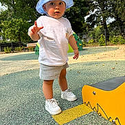 Livio a rejoint le concours — aidez-le/la à gagner de superbes lots ! toddler, child, hat, white_tshirt, shorts, sneakers, playground, outdoor, curious, expression, greenery, trees, bench, fence, sunny, daytime, concrete, play_equipment, young_child, casual_clothing