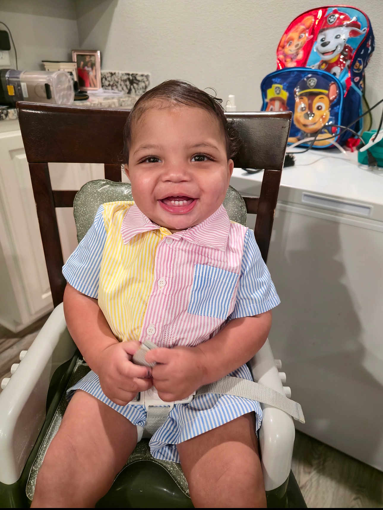 Jahseh is registered to the contest to win money with this photo: toddler, child, high_chair, smiling, indoor, kitchen, colorful_clothing, striped_shirt, baby, happy, seatbelt, furniture, backpack, paw_patrol, counter, cabinet, person, portrait, cute, young_child