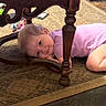 child, girl, toddler, smiling, portrait, lying_down, under_table, wooden_table, ornate_furniture, carpet, indoor, playroom, play_tunnel, colorful, picture_book, toy, pink_shirt, blonde_hair, happy, candid_photo
