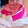 baby, child, face, tongue, pink_clothing, blanket, high_chair, funny_expression, indoor, cute, chubby_cheeks, person, infant, sitting, cozy, soft_texture, wrapped, squinting_eye, casual, portrait