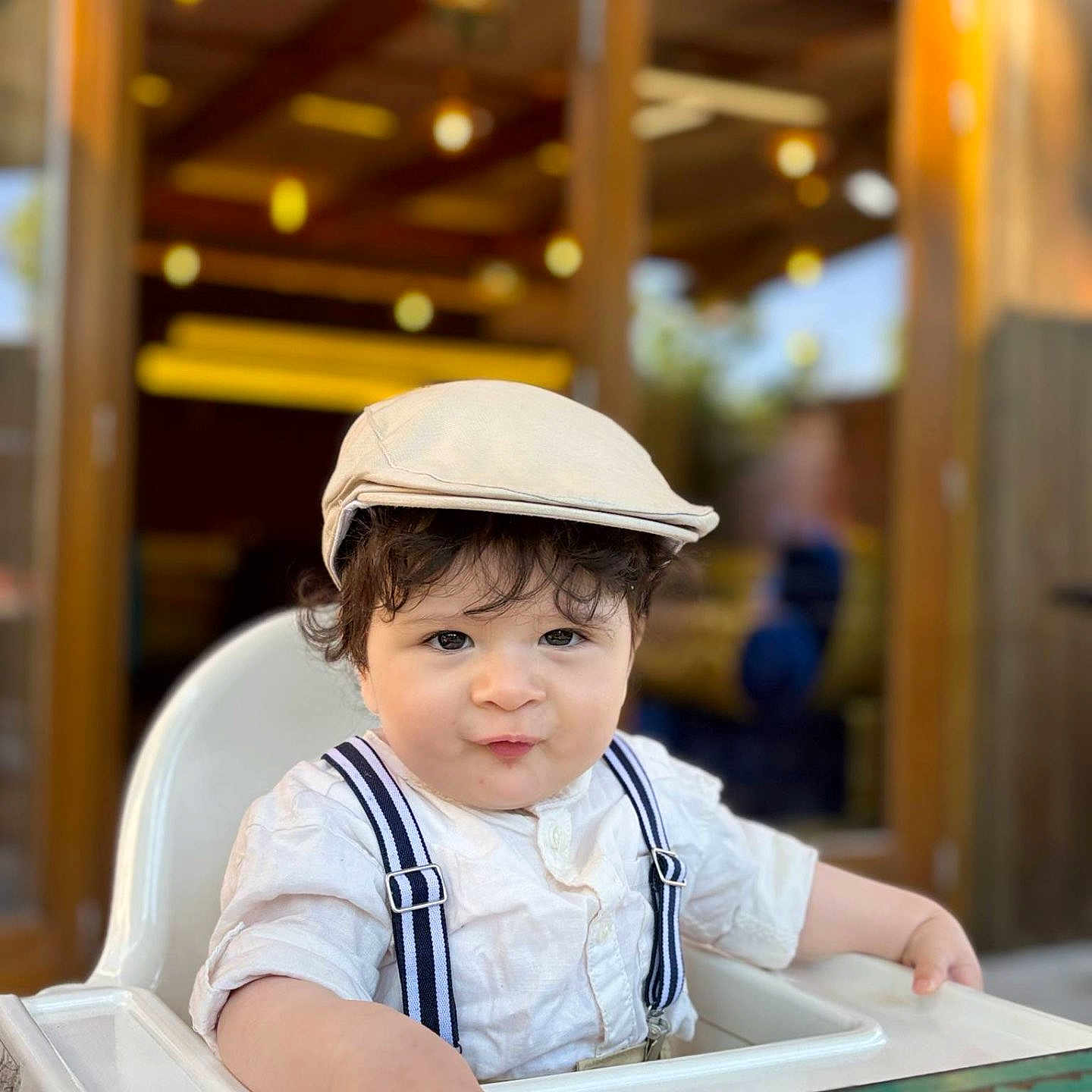 Liam a rejoint le concours — aidez-le/la à gagner de superbes lots ! baby, cap, casual_clothing, child, childhood, curly_hair, cute, face, hat, high_chair, indoor, person, portrait, pout, smile, suspenders, table, toddler, white_shirt, wooden_background