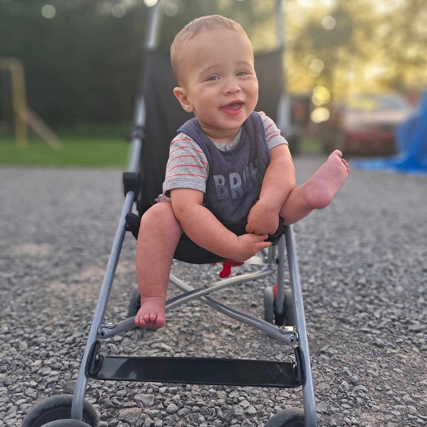 Noah is registered to the contest to win money with this photo: baby, blurred_background, casual_clothing, child, daylight, footwear_absent, gravel, happy, legs, nature, outdoor, park, person, playful, short_sleeves, smiling, stroller, summer, sunlight, toy