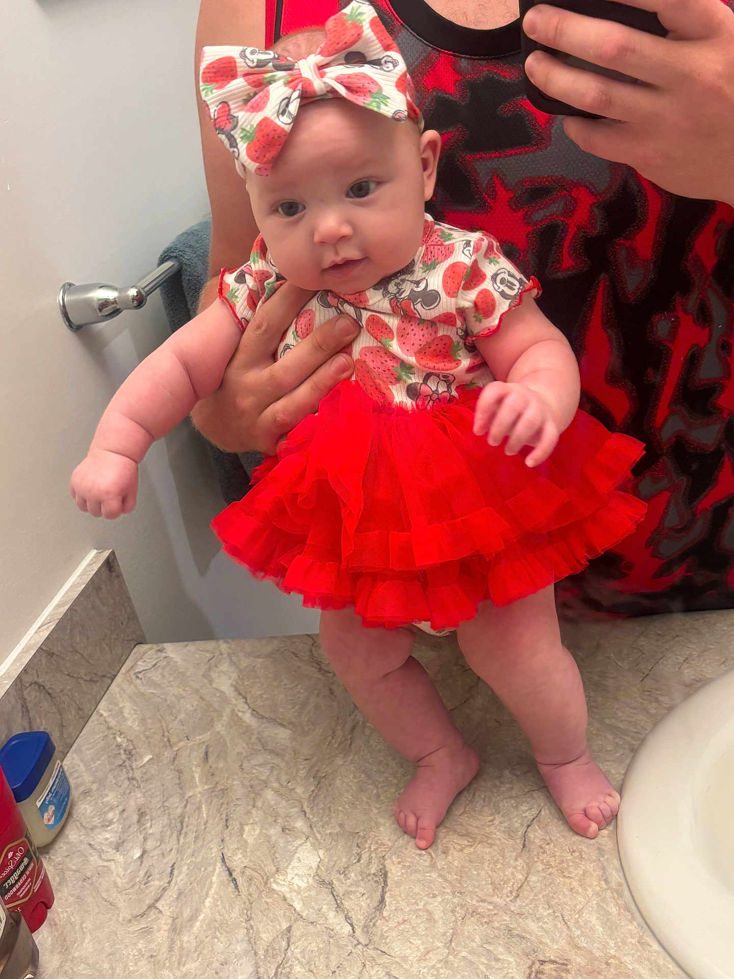 Bradley is registered to the contest to win money with this photo: baby, infant, tutu, red_skirt, headband, strawberry_pattern, mirror, bathroom, sink, towel, hand, reflection, person, clothing, cute, curious, skin, floor, holding, photography