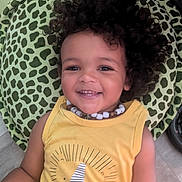 Malik a rejoint le concours — aidez-le/la à gagner de superbes lots ! baby, beaded_necklace, casual_clothing, child, curly_hair, cute, floor, green_cushion, happy, indoor, lion_graphic, patterned_cushion, person, portrait, relaxed, sitting, smiling, toddler, wooden_floor, yellow_tank_top