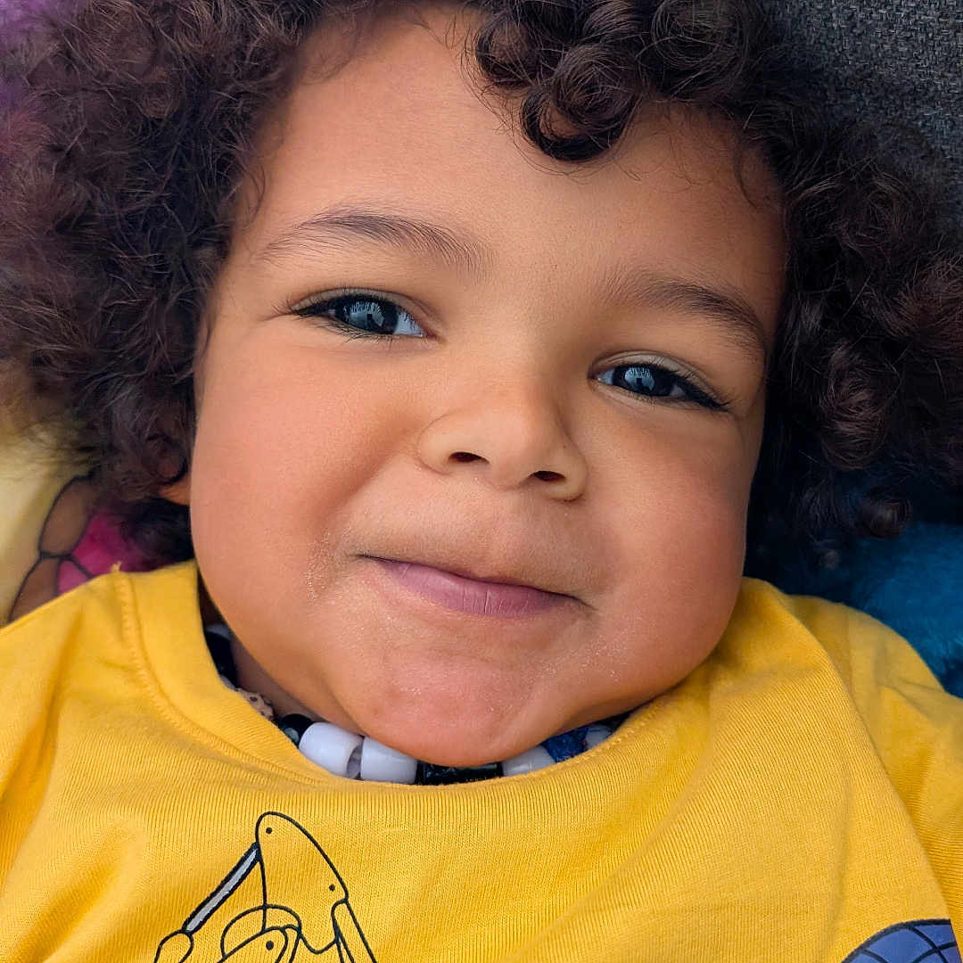 Malik a rejoint le concours — aidez-le/la à gagner de superbes lots ! casual, child, closeup, clothing, construction_vehicle_print, couch, cozy, curly_hair, cute, fabric, face, happy, headshot, indoors, person, portrait, smile, toddler, yellow_shirt, young_child