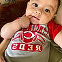 Cruz is registered to the contest to win money with this photo: baby, child, face, cincinnati_reds, shirt, hand, finger_in_mouth, cushion, sofa, indoor, portrait, cute, curious, infant, person, clothing, closeup, resting, young_child, home