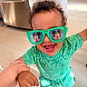bright_colors, casual, child, close_up, curly_hair, dinosaur_pattern, footwear, green_clothing, happy, holding_hand, indoor, joyful, kitchen, person, playful, portrait, smiling, sunglasses, toddler, young_child
