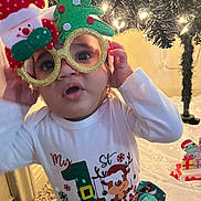 Milo is registered to the contest to win money with this photo: carpet, celebration, child, christmas, clothing, cute, decorations, expression, face, festive, glasses, hand, holiday, indoors, lights, sitting, smiling, toddler, tree, winter