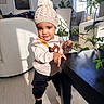 bear_design, black_pants, chair, child, floor, home, indoor, knit_hat, leafy_plant, living_room, person, plant, pom_pom, shadow, socks, sunlight, sweater, table, toddler, window