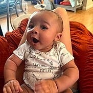 Marley is registered to the contest to win money with this photo: baby, child, infant, onesie, animal_print, smile, happy, person, indoor, furniture, blanket, legs, sitting, face, head, skin, hand, floor, shelf, home