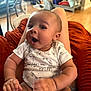 baby, child, infant, onesie, animal_print, smile, happy, person, indoor, furniture, blanket, legs, sitting, face, head, skin, hand, floor, shelf, home
