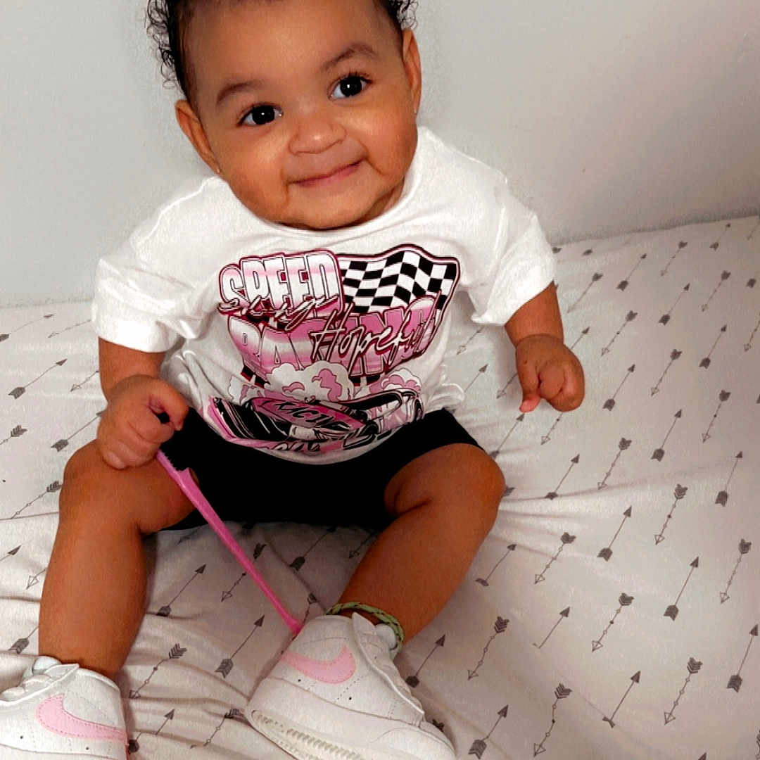 Mahala joined the competition — help win amazing prizes! arrows_pattern, bedspread, black_shorts, child, curly_hair, cute, footwear, graphic_tee, happy, indoor, person, pink_accents, playing, shoelace, sitting, smiling, toddler, white_sneakers, white_tshirt, young