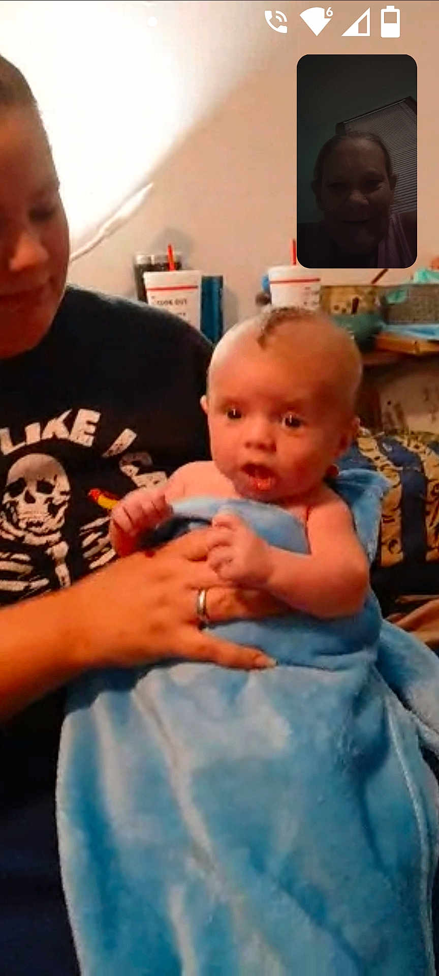 Tony is registered to the contest to win money with this photo: baby, child, adult, blanket, blue_blanket, holding, indoor, video_call, screen_inset, smile, person, face, hand, clothing, shirt, skeleton_design, furniture, room, window, curtain