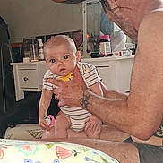 Kodi is registered to the contest to win money with this photo: baby, man, tattoo, indoor, furniture, guitar, pacifier, person, holding, striped_clothing, cabinet, bottle, mirror, face, hand, sitting, curious, skin, adult, room
