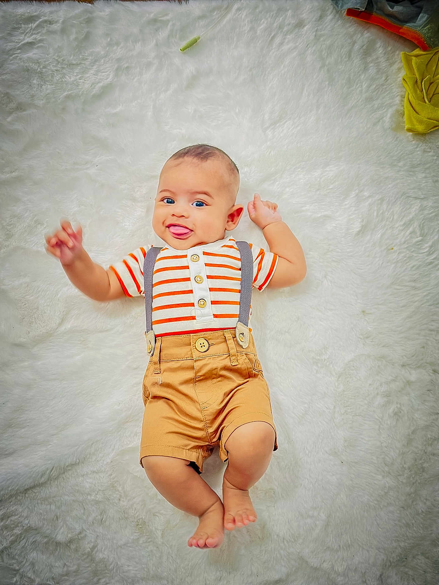 Thabit is registered to the contest to win money with this photo: baby, child, infant, smiling, tongue_out, striped_shirt, suspenders, shorts, barefoot, lying_down, white_rug, happy, cute, playful, indoor, portrait, person, small_child, funny_face, casual_clothing