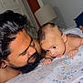 baby, man, bed, floral_bedspread, face, beard, curly_hair, indoor, room, lamp, door, skin, close_up, portrait, interaction, parent_and_child, tender_moment, lying_down, expression, looking